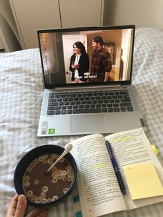 a laptop computer sitting on top of a bed next to a bowl of cereal and an open book