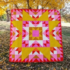 a pink and yellow quilt sitting on top of a leaf covered ground next to a tree