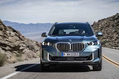 the front end of a silver bmw x5 driving down a mountain road