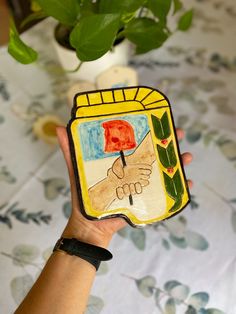 a person holding up a small painting in their hand with a flower pot behind it