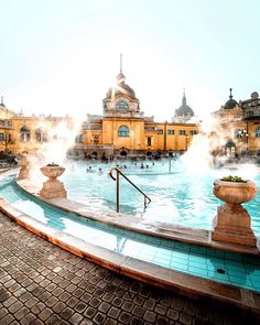 an outdoor swimming pool with fountains and people in the water near buildings on either side