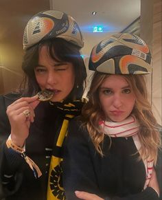 two young women wearing hats and scarves eating food from a spoon in front of them