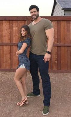 a man and woman standing next to each other in front of a wooden fence with their arms around each other