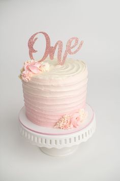 a white cake with pink frosting and flowers on top that says one in the middle