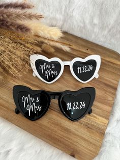 two heart shaped sunglasses with the names of each couple and date on them, sitting on top of a wooden board