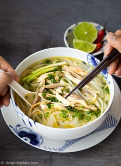 two hands holding chopsticks over a bowl of chicken noodle soup with lemon and cilantro