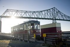 a red and white train traveling down tracks under a large metal bridge with a person standing on it