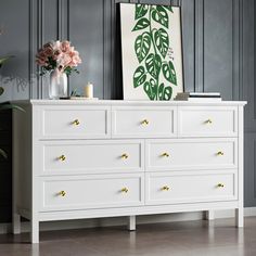 a white dresser with gold handles and drawers in a living room next to a painting