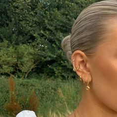 a woman with blonde hair wearing gold earrings and a white dress in front of trees