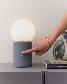 a person pointing at a lamp on top of a table next to books and a vase