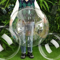 a man standing in front of two clear bubble balls on green grass with trees and bushes behind him