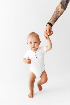 a baby in a white bodysuit holding the hand of an adult's hand