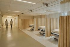 two people walking down a hospital hallway with beds and curtains hanging from the ceiling in front of them