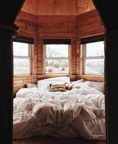 an unmade bed sitting in the middle of a room next to two large windows