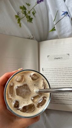 a person is holding a drink in their hand and an open book on the bed