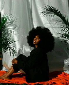 a woman with an afro sitting on the ground in front of a white curtain and palm trees