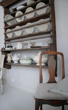 an old wooden shelf filled with dishes on top of a white wall next to a chair