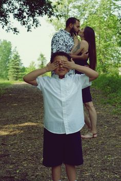 a man covering his eyes while standing next to a woman on a dirt road with trees in the background