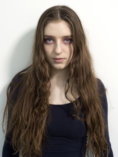 a woman with long brown hair and blue eyes posing for a photo in front of a white wall