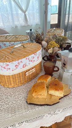 bread and coffee on a table in front of a window with curtains, flowers and other items