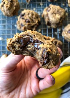 a person holding up a cookie in front of a banana and other cookies on a cooling rack