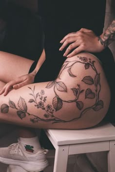 a woman sitting on top of a white stool next to a chair with tattoos on her legs