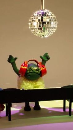 a green and red stuffed animal sitting on top of a bench next to a disco ball