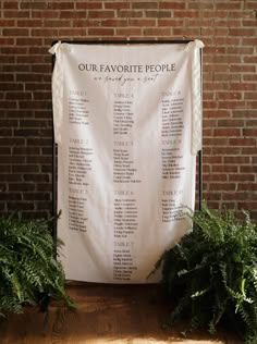 a white sign sitting on top of a wooden floor next to green plants and a brick wall