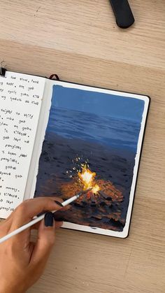 a person holding a pencil in their hand and drawing on an open notebook with a fire