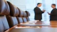 two men in suits are standing behind a conference table with papers and pens on it