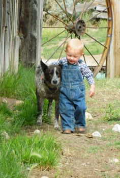 a young boy standing next to a dog in the grass near a wooden structure with a spoke wheel behind him