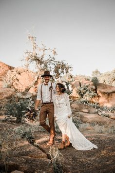 a bride and groom walking through the desert together in their wedding attire, holding hands