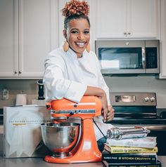 a woman standing in front of a kitchen counter with an orange mixer and books on the counter