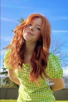 a woman with long red hair wearing a green and white checkered dress standing in front of a fence