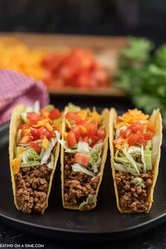 three tacos on a black plate with lettuce and tomatoes in the background