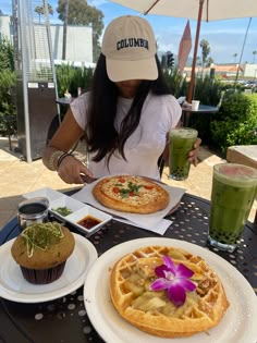 a woman sitting at an outdoor table with two plates of food and drinks in front of her