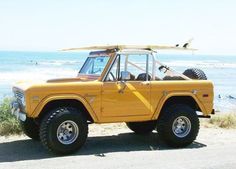 an old yellow jeep with a surfboard on top parked in front of the ocean