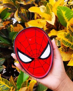 a hand holding a red and black spider - man coaster in front of green leaves