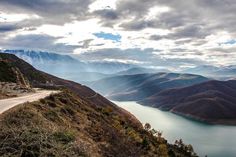 a scenic view of mountains and water from the top of a hill with clouds in the sky