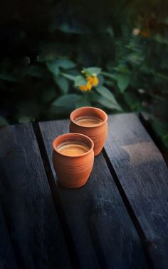 two clay cups filled with liquid sit on a wooden table in front of some flowers