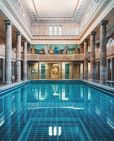 an indoor swimming pool in a large building with columns on either side and people sitting at the top