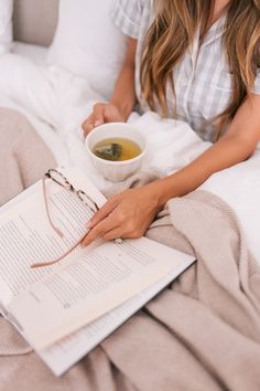 a woman sitting in bed reading a book and holding a cup of tea next to her