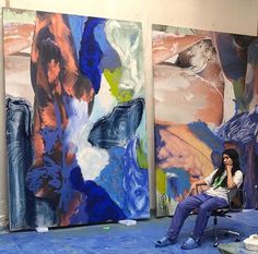 a woman is sitting on a bench in front of two large abstract paintings that are being worked on