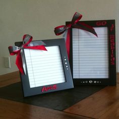 two black frames with red ribbon and writing paper attached to them, sitting on top of a wooden table