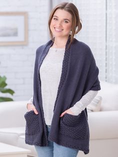 a woman standing in front of a white couch wearing a blue cardigan sweater and jeans