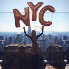 a woman standing on top of a tall building holding up giant gold letter balloons in the air