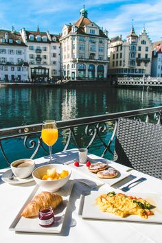 an outdoor table with food and drinks on it near the water's edge in front of some buildings