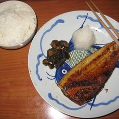 a white plate topped with fish next to rice and chopsticks on top of a wooden table