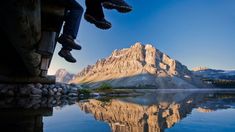 a person sitting on the edge of a dock next to a mountain lake with mountains in the background