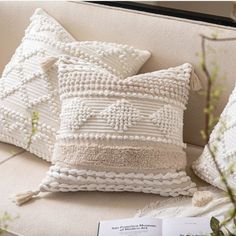 three white pillows sitting on top of a couch next to a book and planter